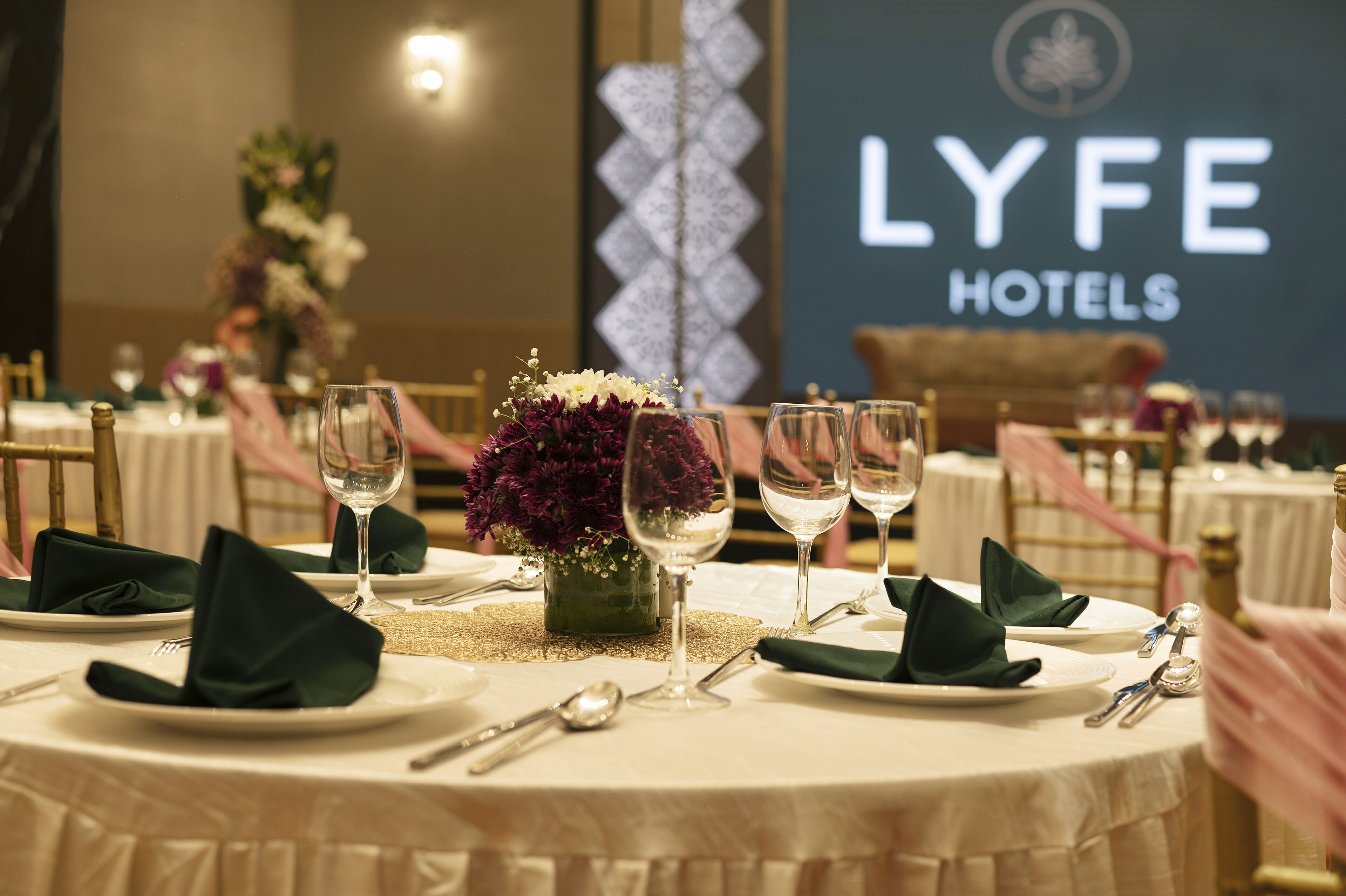 A table set for a formal dining experience with a floral centerpiece, silverware, and glassware, in a room with a 'LYFE HOTELS' sign in the background.