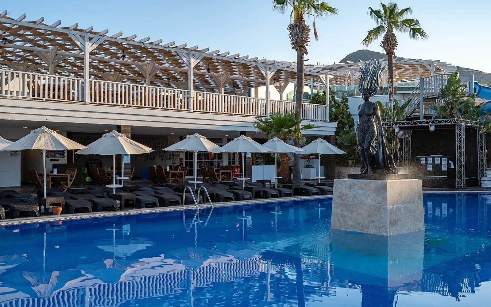Zona de piscina amb gandules i ombrel·les, una estàtua i una zona de seients coberta amb vista a les muntanyes.
