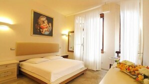 Ein Hotelzimmer mit einem großen Bett, einem Nachttisch mit Blumenvase, einem Bild an der Wand und einem Fenster mit Vorhängen.