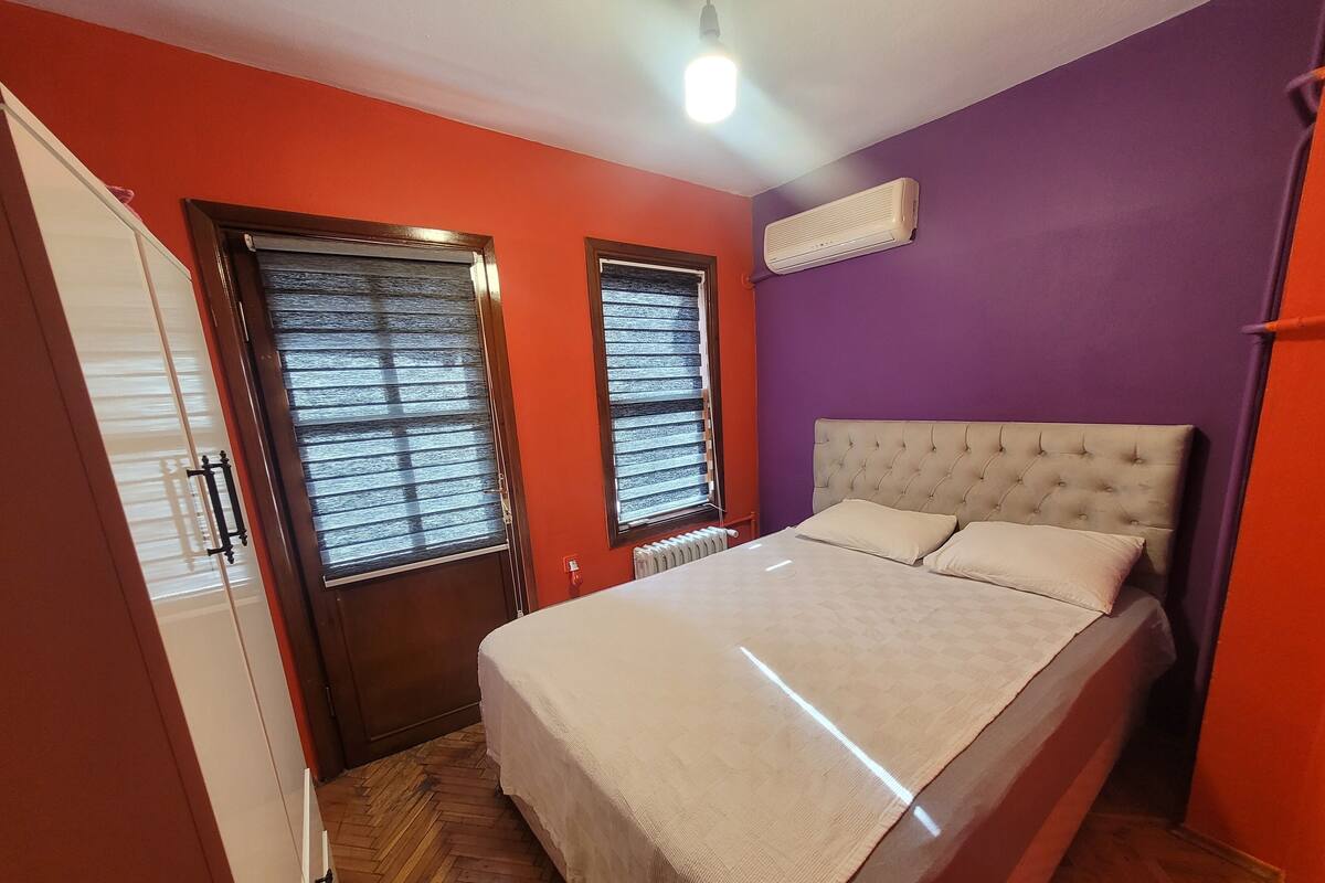 A bedroom with a bed, two windows with blinds, a radiator, and a ceiling light.