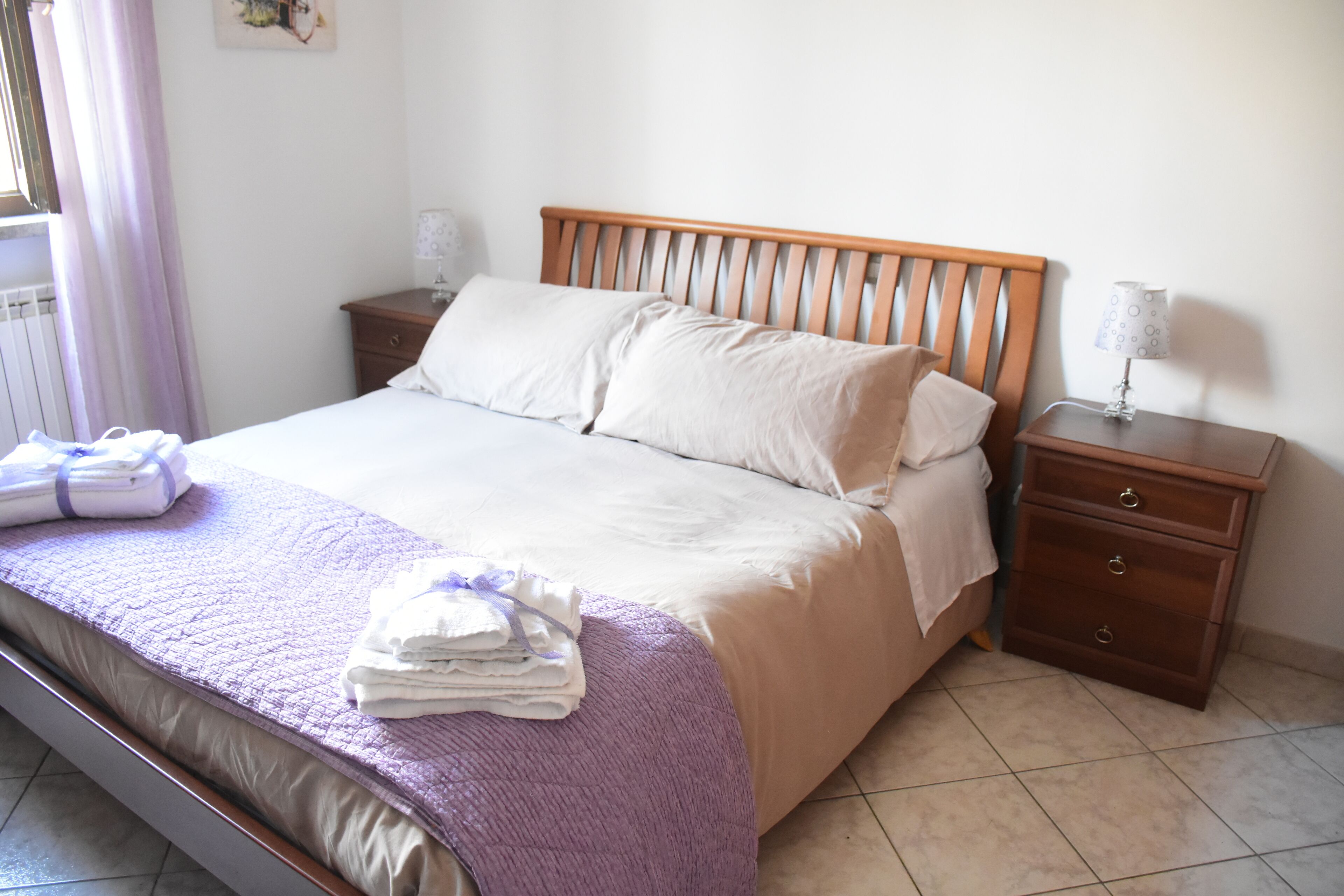 A neatly made bed with a wooden headboard, a bedside table with a lamp, and a stack of folded towels on the floor.