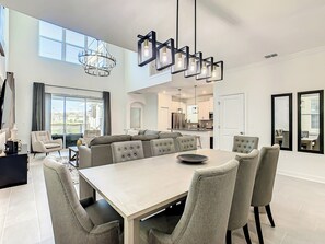 A modern dining room with a large table and chairs, a chandelier, and a view of the kitchen and living area.