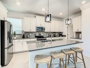 A modern kitchen with white cabinetry, stainless steel appliances, a central island with stools, and pendant lighting.