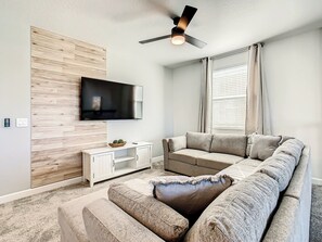 Un salon moderne doté d’un téléviseur à écran plat fixé à un mur en bois, d’un canapé d’angle, d’un meuble de rangement blanc et d’un ventilateur de plafond.