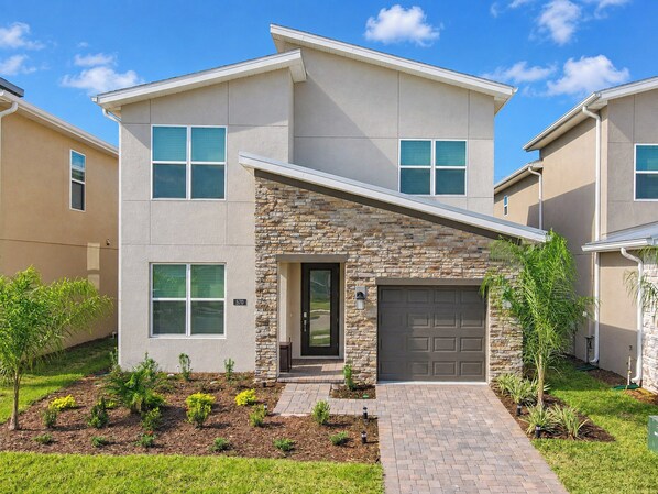 A modern two-story residential building with a stone facade, a front garage, and a landscaped yard with small trees and shrubs.
