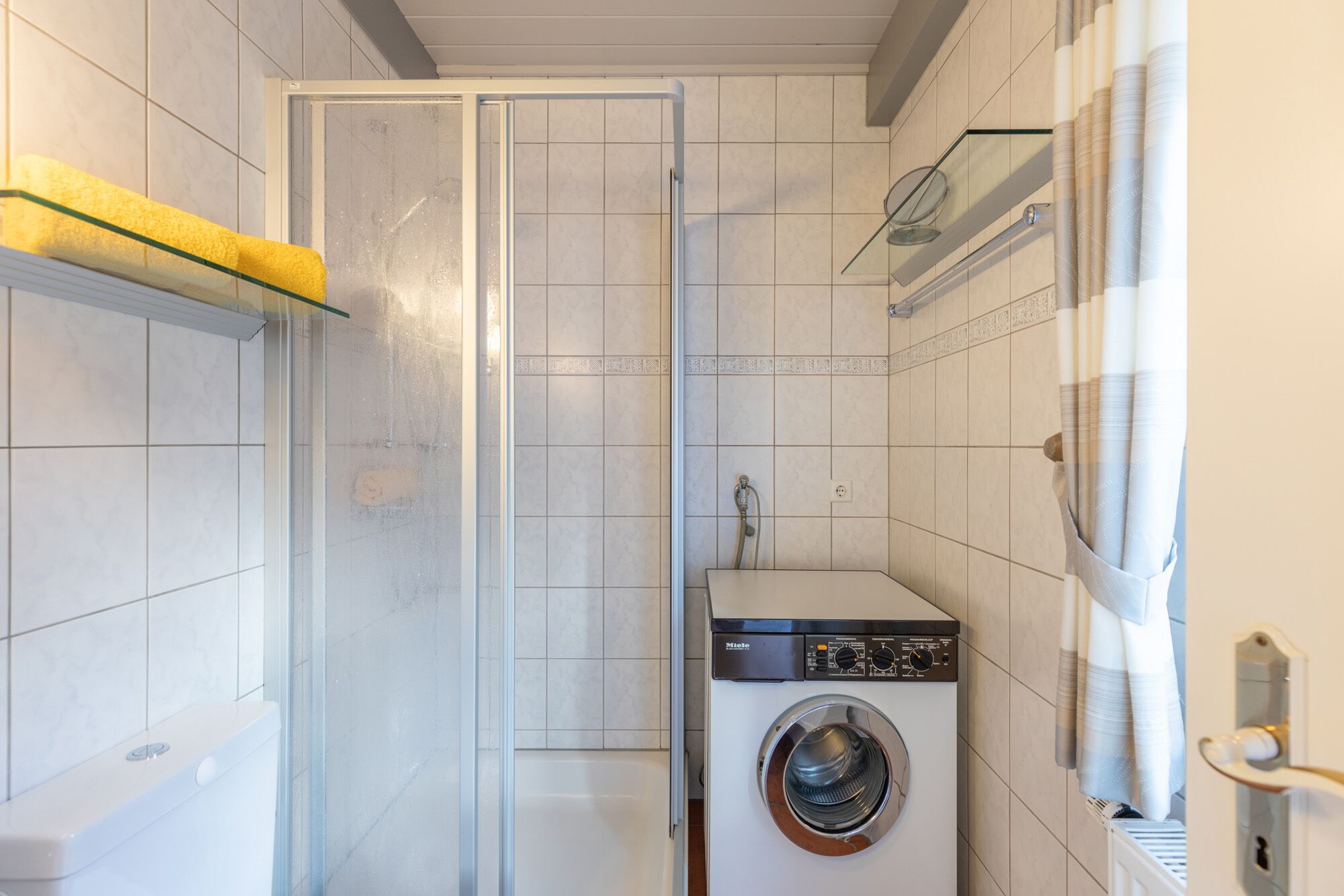 A compact bathroom with a glass shower enclosure, a washing machine, and a toilet.