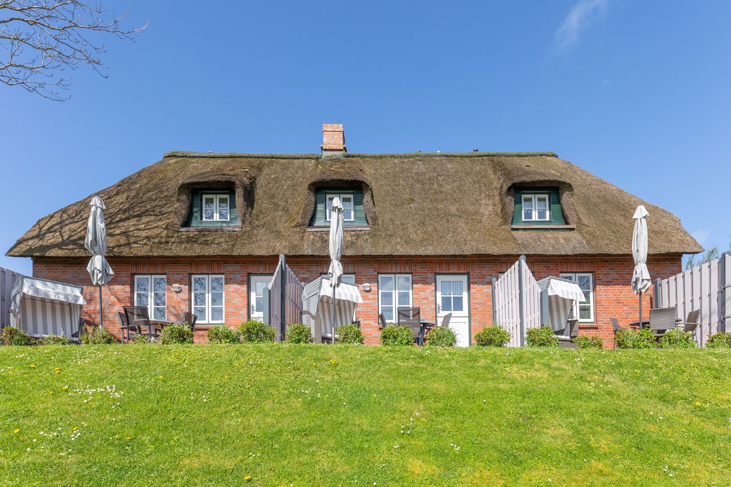Une maison traditionnelle en briques, avec un toit de chaume, de nombreuses fenêtres et du mobilier d’extérieur.