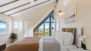 Quarto com cama grande, mesa de cabeceira, planta em vaso e vista do exterior pela janela.