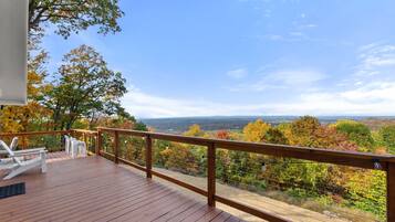 Une terrasse en bois, agrémentée de chaises blanches et d’une balustrade, offre une vue panoramique sur le paysage automnal et les collines lointaines.