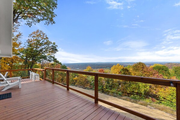 Una terraza de madera con sillas blancas y barandilla, con vistas panorámicas al follaje otoñal y colinas distantes.