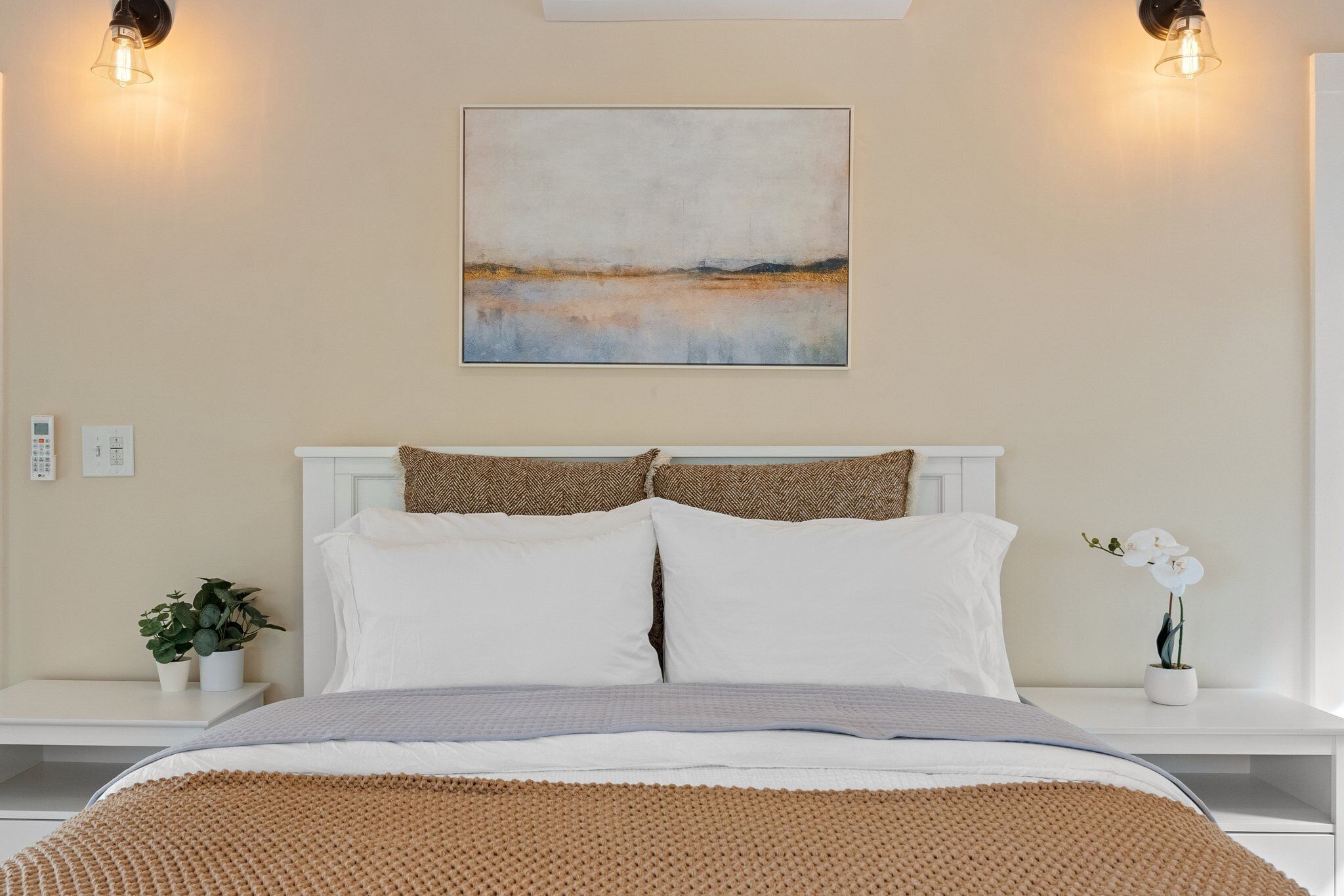 Un dormitorio con cama, ropa de cama blanca, un cuadro en la pared y mesitas de noche con plantas.