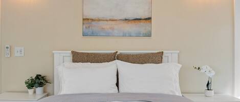 Un dormitorio con cama, ropa de cama blanca, un cuadro en la pared y mesitas de noche con plantas.