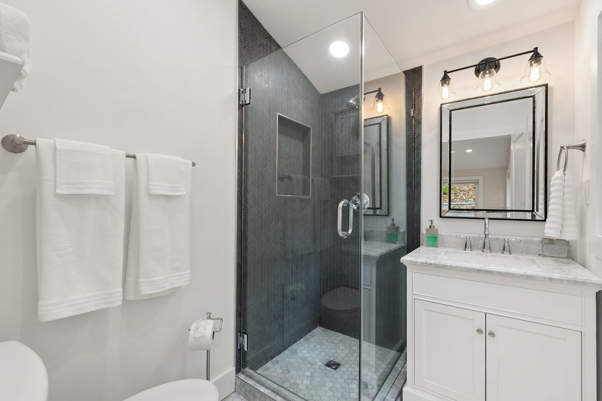Een moderne badkamer met een douchecabine van glas, een wit wastafelmeubel met een grote spiegel, en twee witte handdoeken aan de muur.