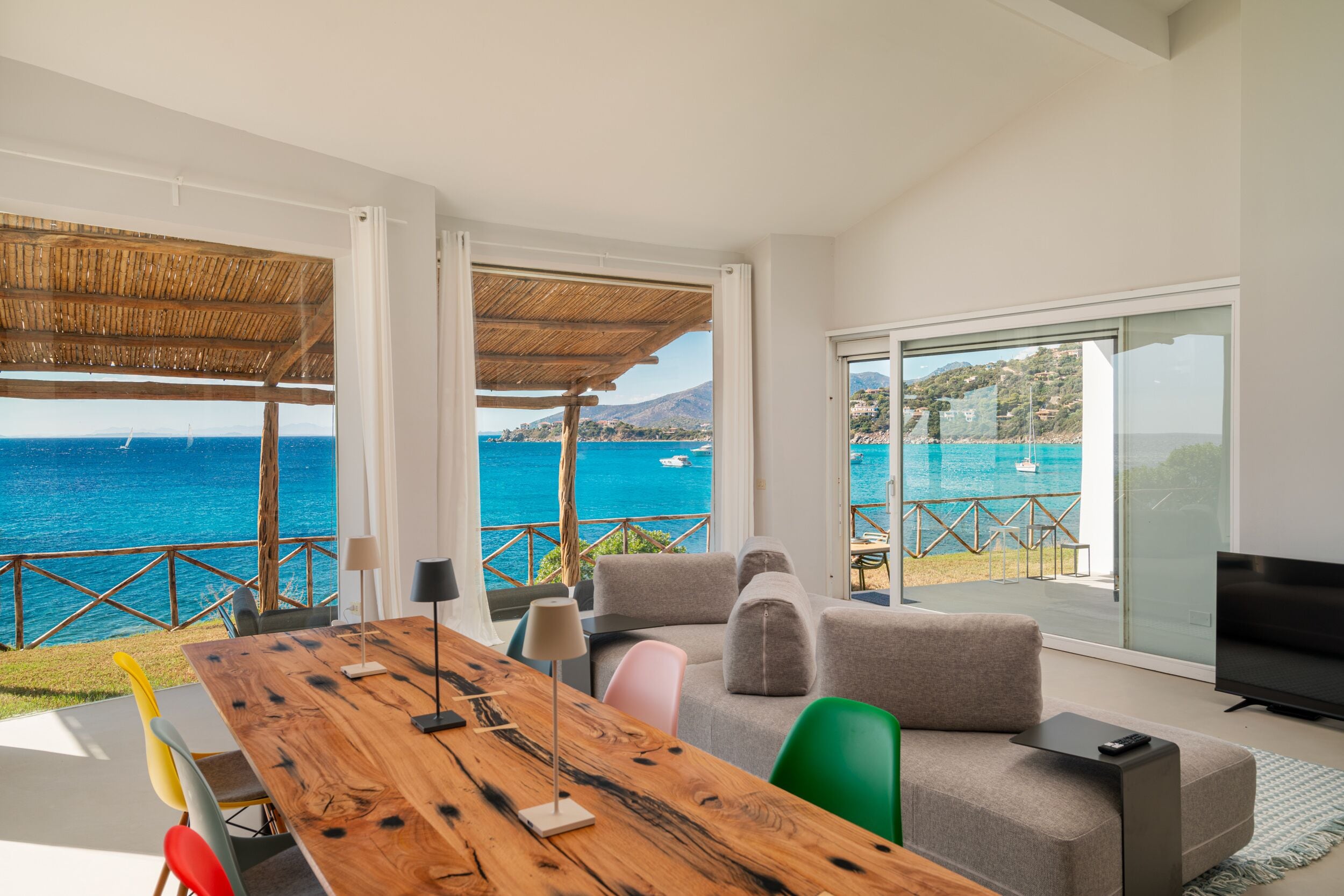 A modern living room with a wooden dining table, grey sofas, and a large TV. It features a balcony with a view of the sea and a mountain.