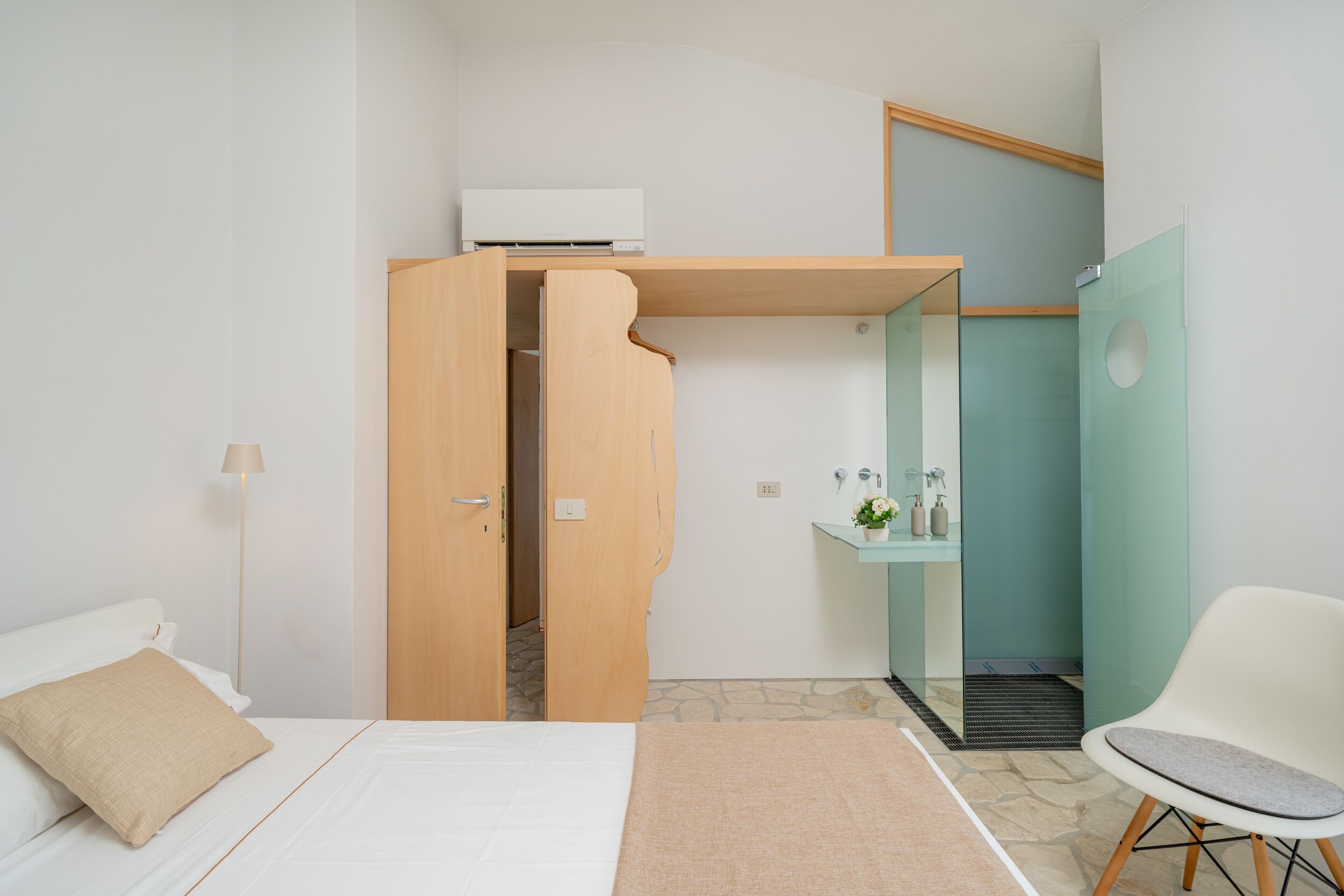 A hotel room with a bed, a wooden wardrobe, a bathroom with a glass shower, and a white chair.