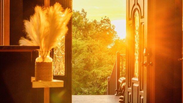 Uma porta aberta dá para uma varanda com vista para árvores e um pôr do sol.