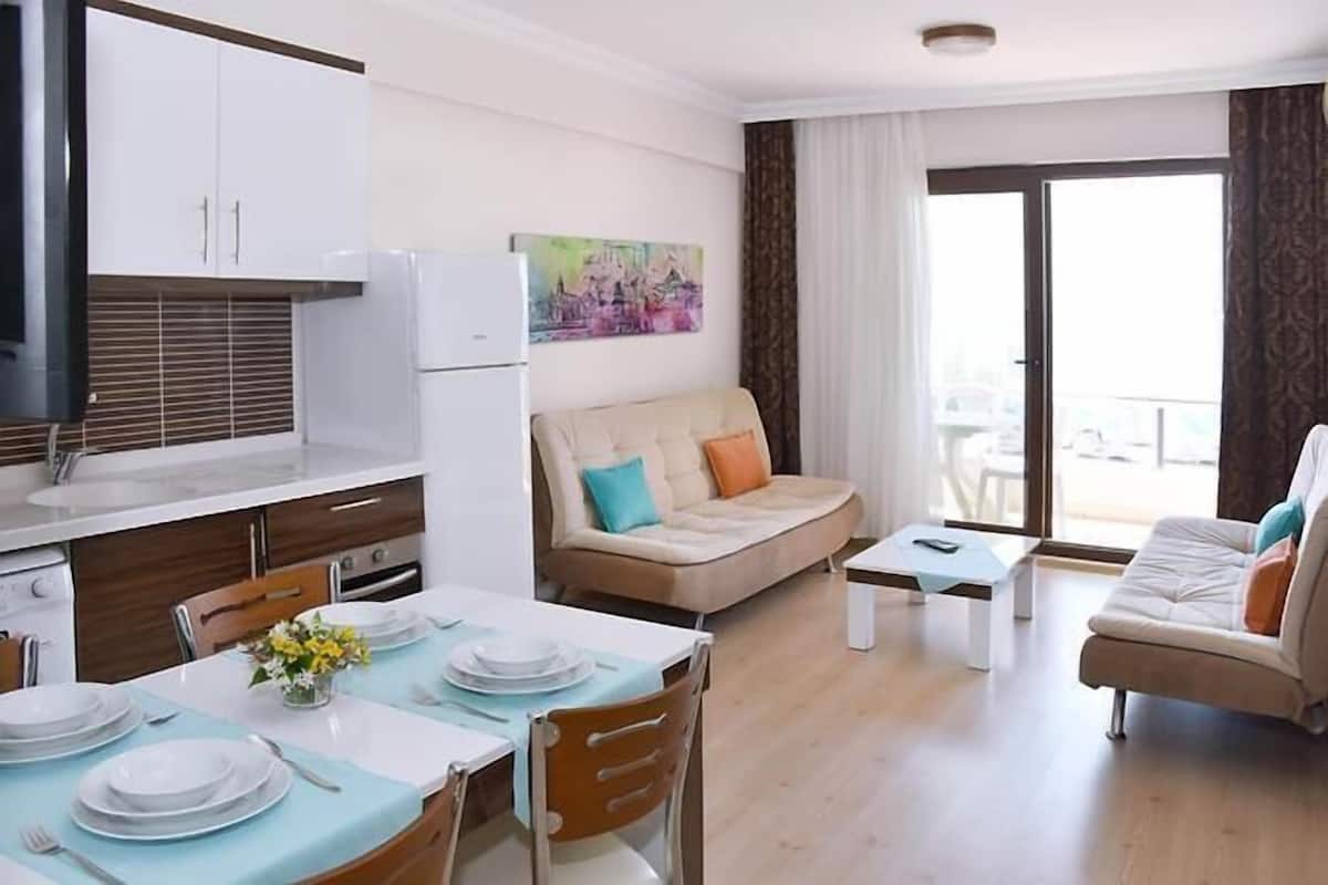 A modern living room with a sofa, a dining table set with plates, and a kitchen area with cabinets and a refrigerator.