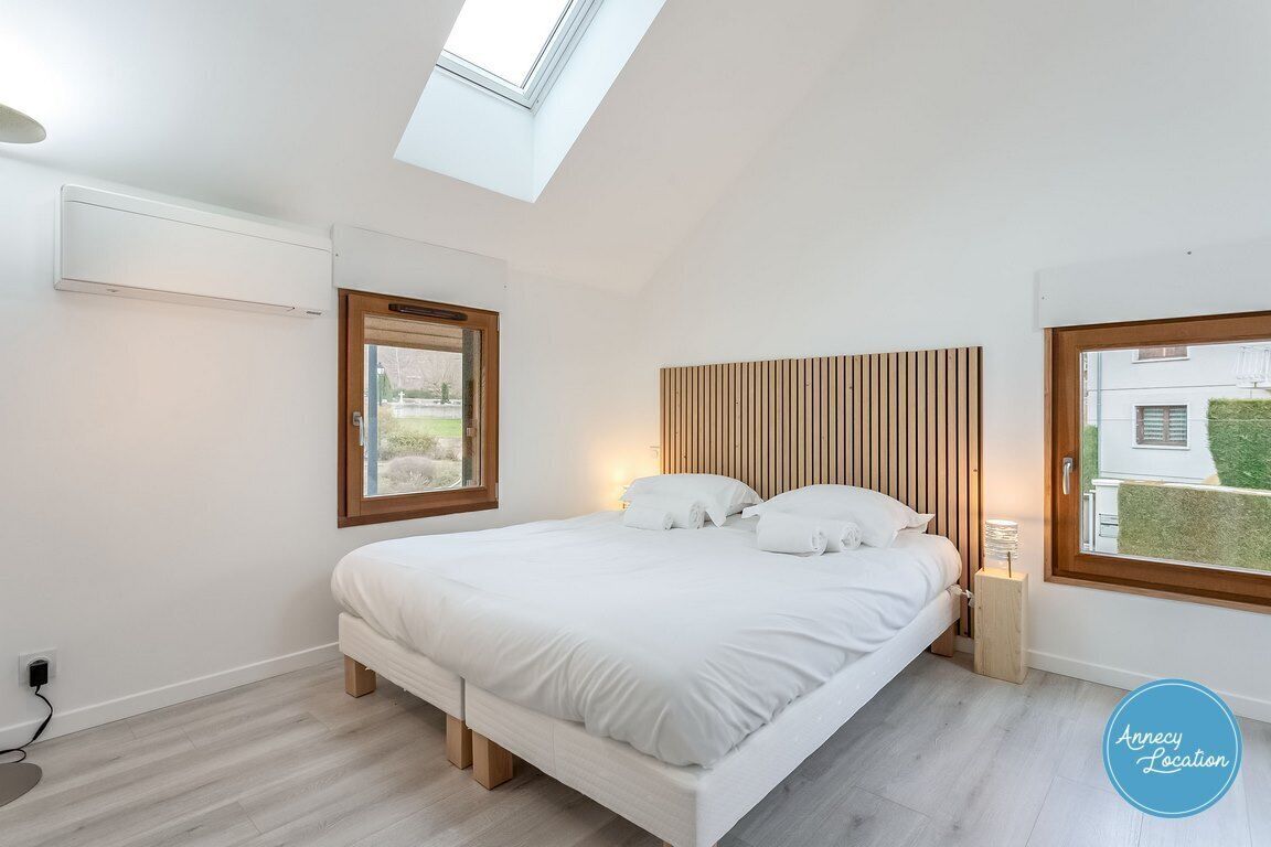 Ein modernes Schlafzimmer mit Dachfenster, einem Bett mit weißer Bettwäsche, einem Holz-Kopfende und Blick auf ein Haus durch ein Fenster.