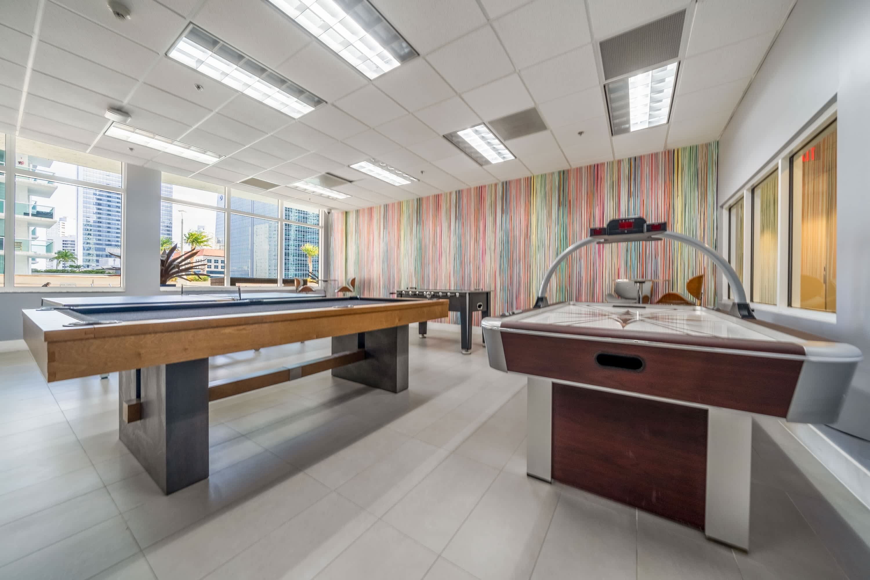Un espace piscine intérieur moderne, doté de deux tables de billard, de grandes fenêtres et d’un mur aux rayures colorées.