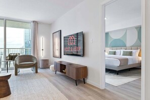 Une chambre d’hôtel moderne dotée d’un grand lit, d’un coin salon avec un fauteuil et une petite table, d’une console et d’une grande fenêtre donnant sur la ville.