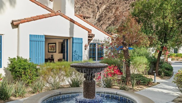 Un bâtiment aux volets bleus, une fontaine et un paysage montagneux en arrière-plan.