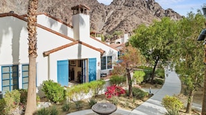 A white building with blue shutters, a stone fountain, and a mountain backdrop.