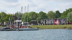 A marina with several sailboats docked, a restaurant with outdoor seating, and a grassy area with trees in the background. - Floating 44 Komfortable Hausboote bei Xanten mit Tollem Seeblick (Xanten)