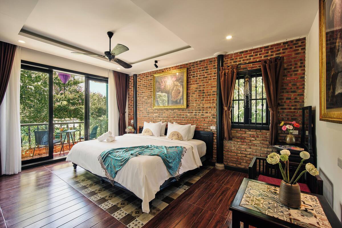 Une chambre avec un grand lit, un ventilateur de plafond, un mur en briques et une fenêtre avec des rideaux.