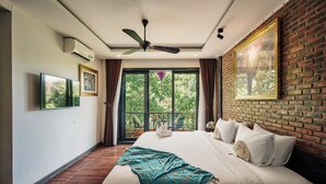 Habitación de hotel con una cama grande, ventilador de techo, televisor y vistas a zonas verdes a través de las ventanas.