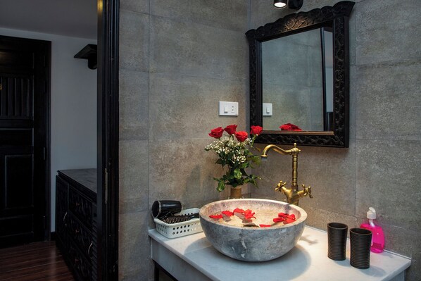 Un baño con lavamanos de piedra, un espejo, un florero con rosas rojas y un grifo dorado.