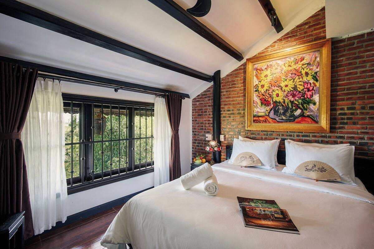 Une chambre d’hôtel avec un grand lit, une fenêtre avec des rideaux, un mur en briques et un tableau représentant des fleurs.