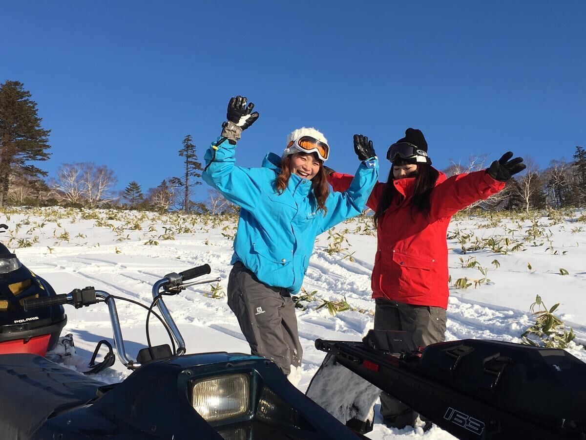 Two people in winter clothing posing with snowmobiles in a snowy field.