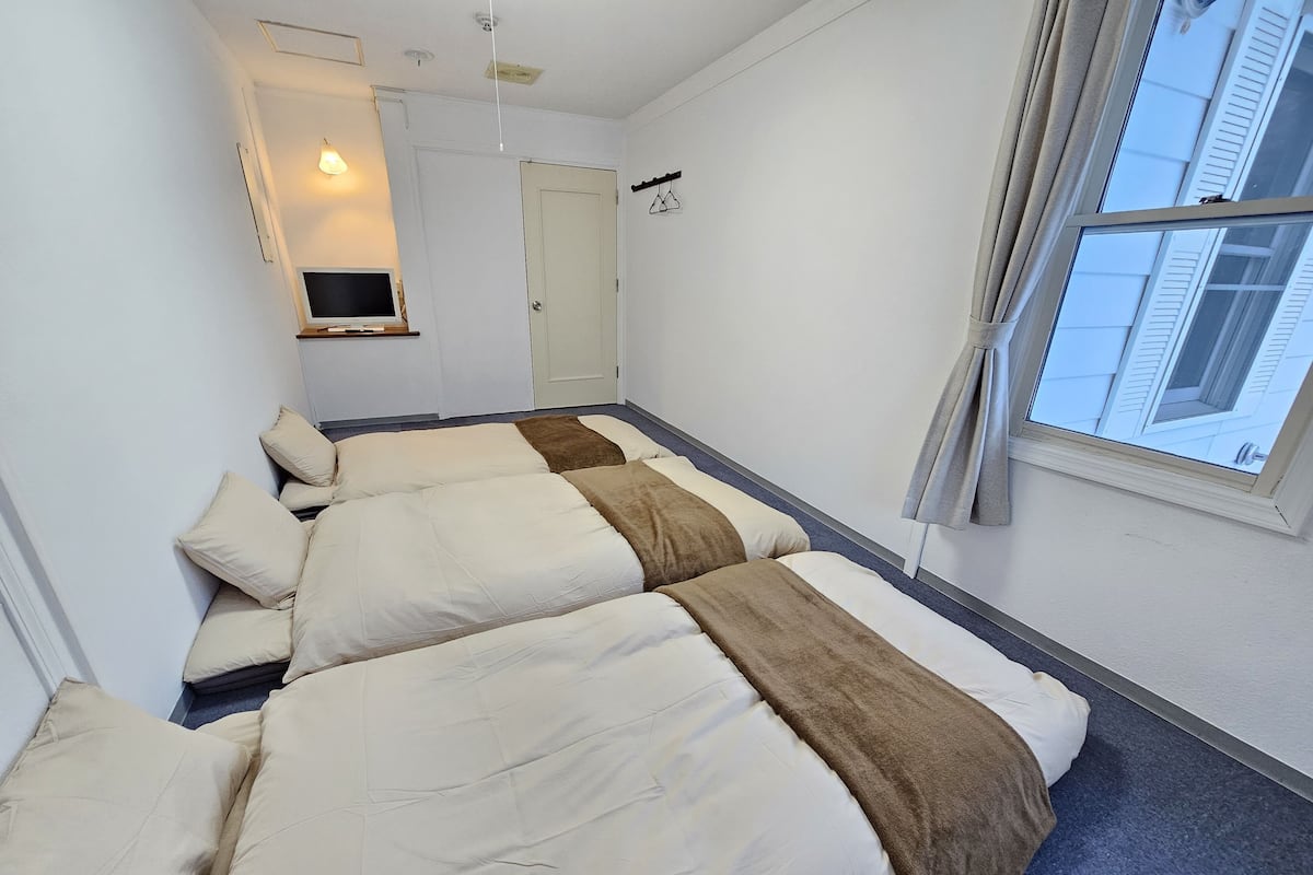 A room with three beds, a television, a door, and a window with a view of buildings.