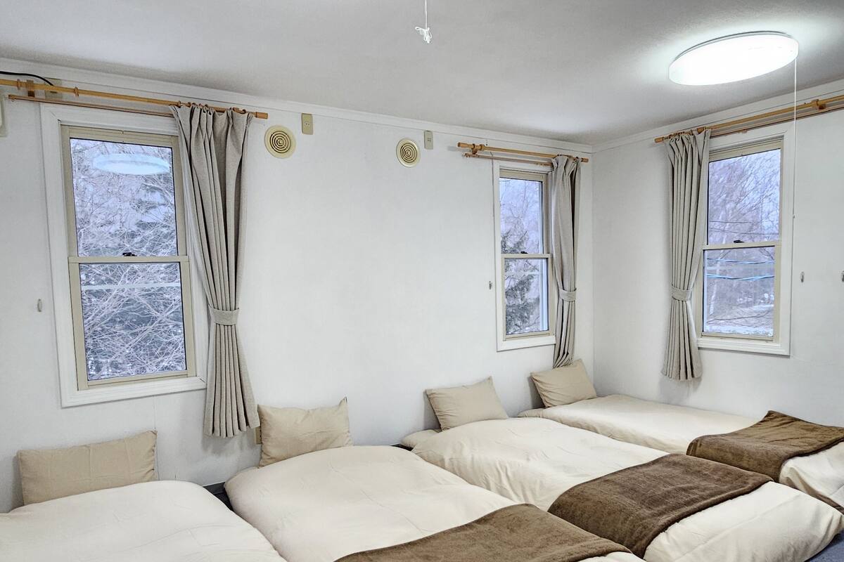 A room with four beds arranged in a U-shape, large windows with curtains, and a ceiling light.