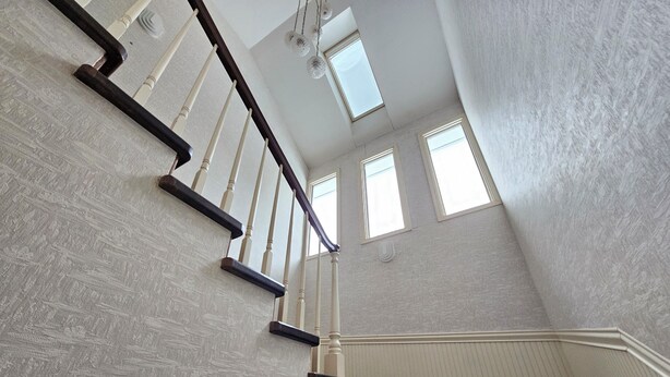 Una escalera con pared texturizada y barandilla de madera, que conduce a un rellano con vista al cielo a través de un tragaluz.