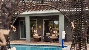 Un espace piscine avec des fauteuils en osier et un canapé, un bâtiment doté d’un toit en tuiles, et une personne debout près de la piscine.