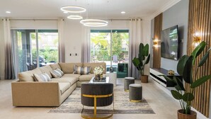 Un salon moderne avec un canapé, une table basse et un téléviseur. On peut voir des plantes, un tapis et un balcon.