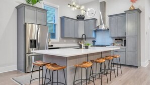A modern kitchen with grey cabinets, a stainless steel refrigerator, a large island with bar stools, and a pendant light. - New Home Great for Groups | Heated Pool, Sleeps 10 (New Orleans)