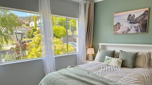 A bedroom with a large window, a bed with a green and white striped bedspread, a painting of a coastal scene, and a lamp on a bedside table. - SandyBay Garden Hideaway (Sandy Bay)