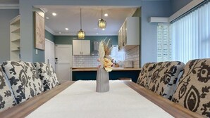 A dining area with a long table, patterned cushions, a vase with flowers, pendant lights, and a kitchen in the background. - SandyBay Garden Hideaway (Sandy Bay)