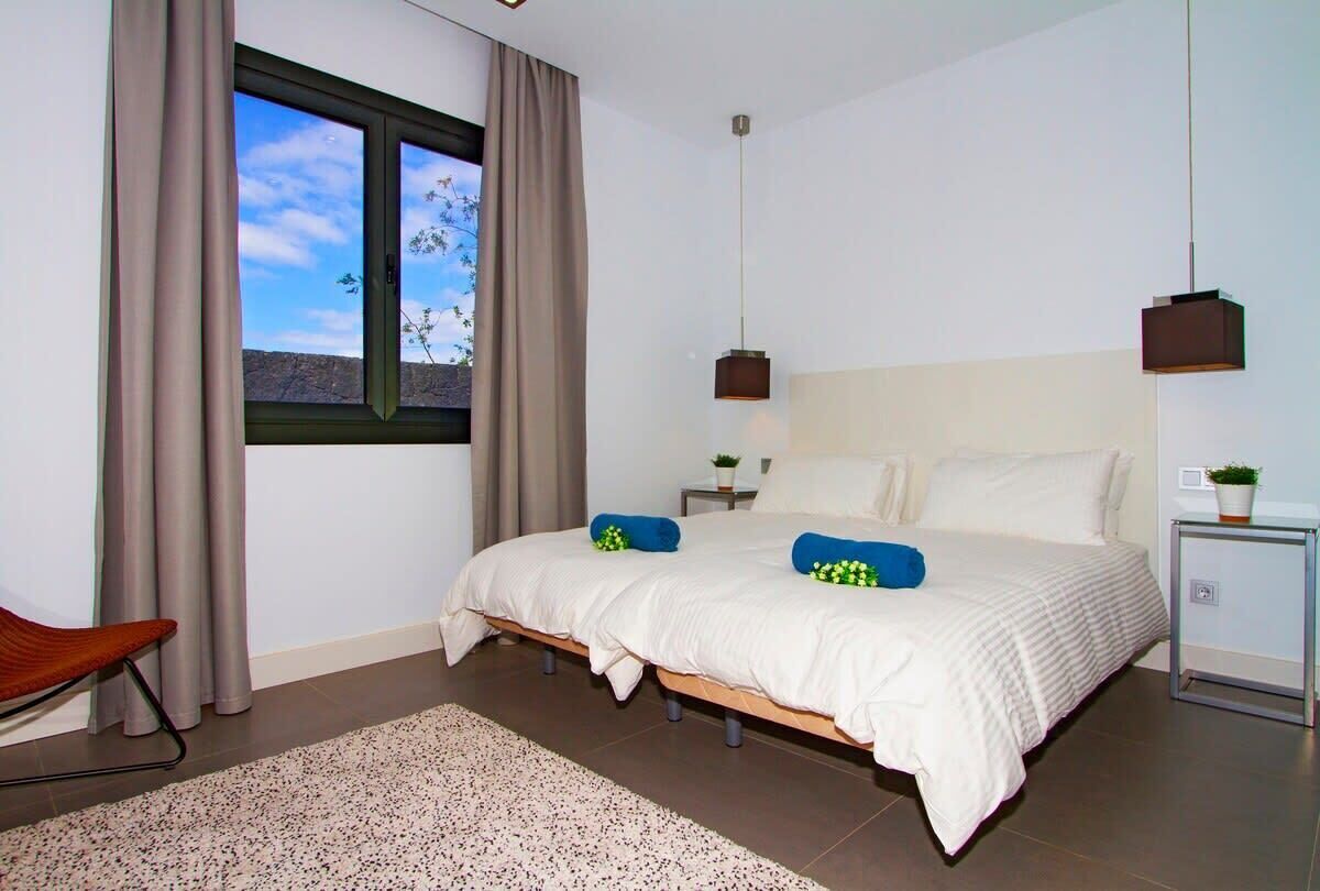 Un dormitorio con una cama grande, una ventana con cortinas, una mesita de noche con una lámpara y una planta pequeña.