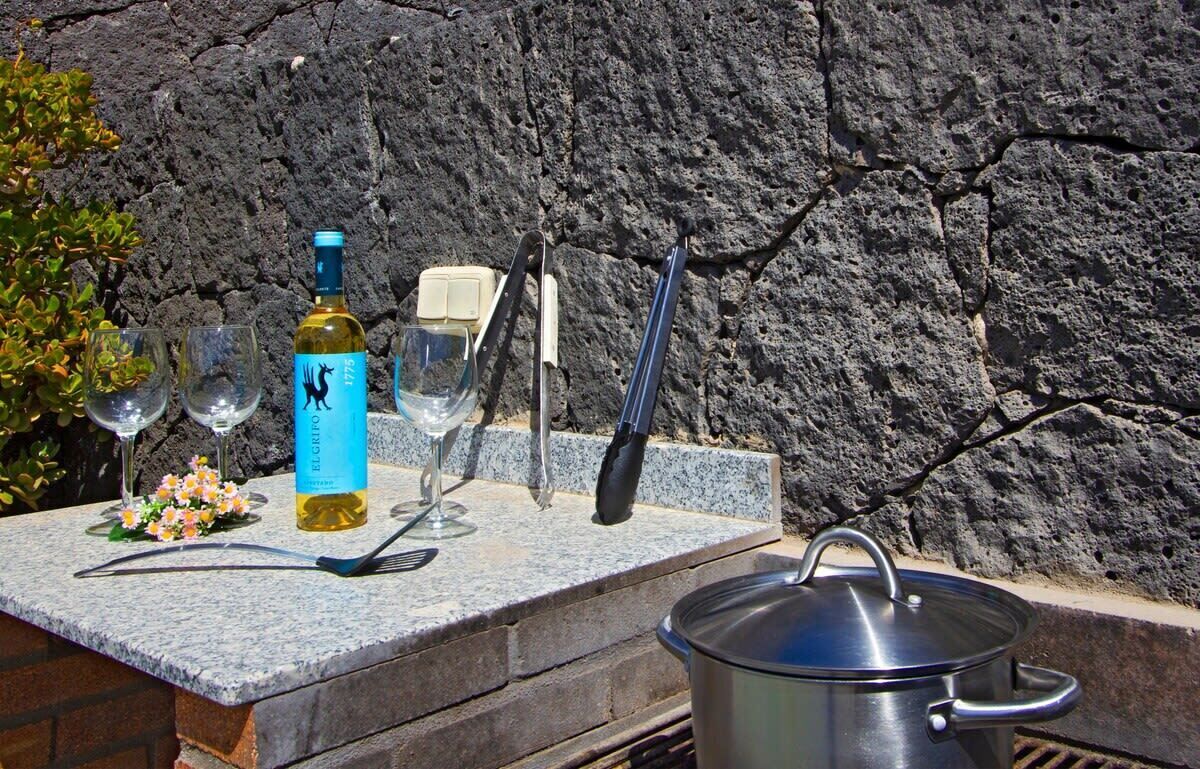 A table with a bottle, wine glasses, a pot, and cooking utensils against a rocky wall.