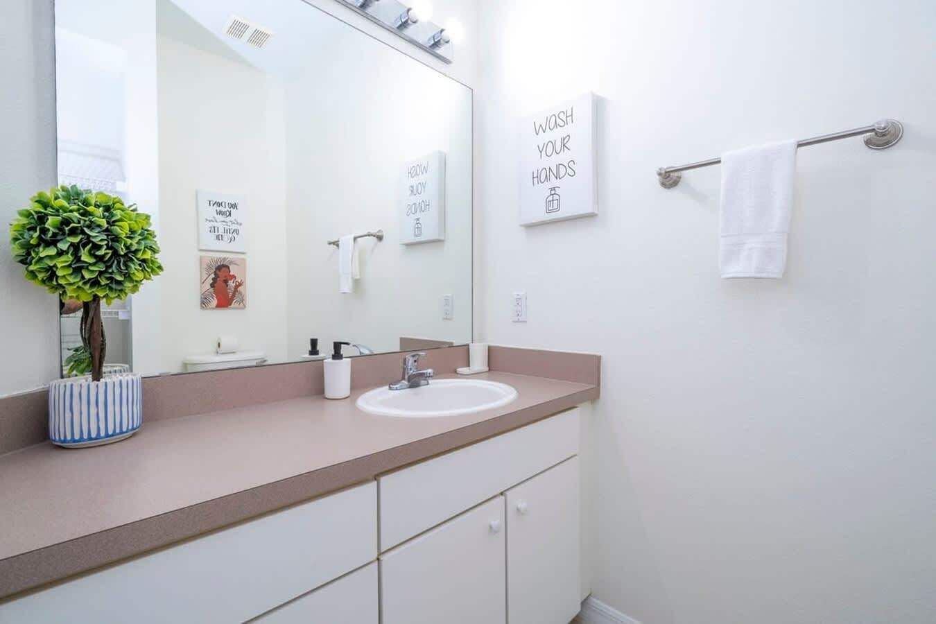 Un baño moderno con un lavamanos blanco, un espejo grande, un perchero para toallas y un cartel que dice 'LÁVATE LAS MANOS'.