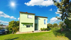 A two-story house with a green facade and a red-tiled roof, surrounded by a well-maintained lawn and trees. - Holiday apartment with air conditioning (Banjole)