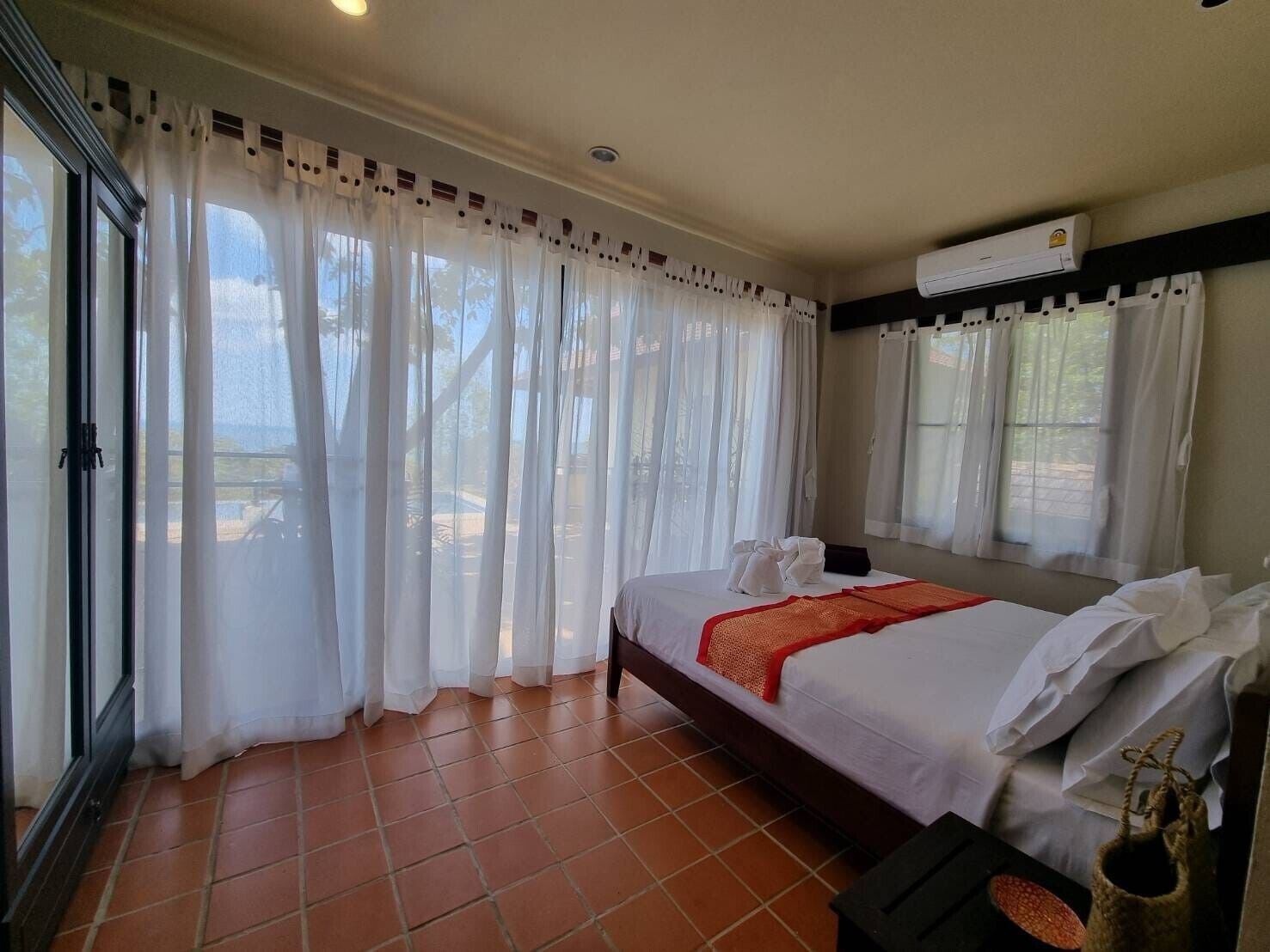 A bedroom with a bed, white curtains, an air conditioning unit, and a view of the outdoors.