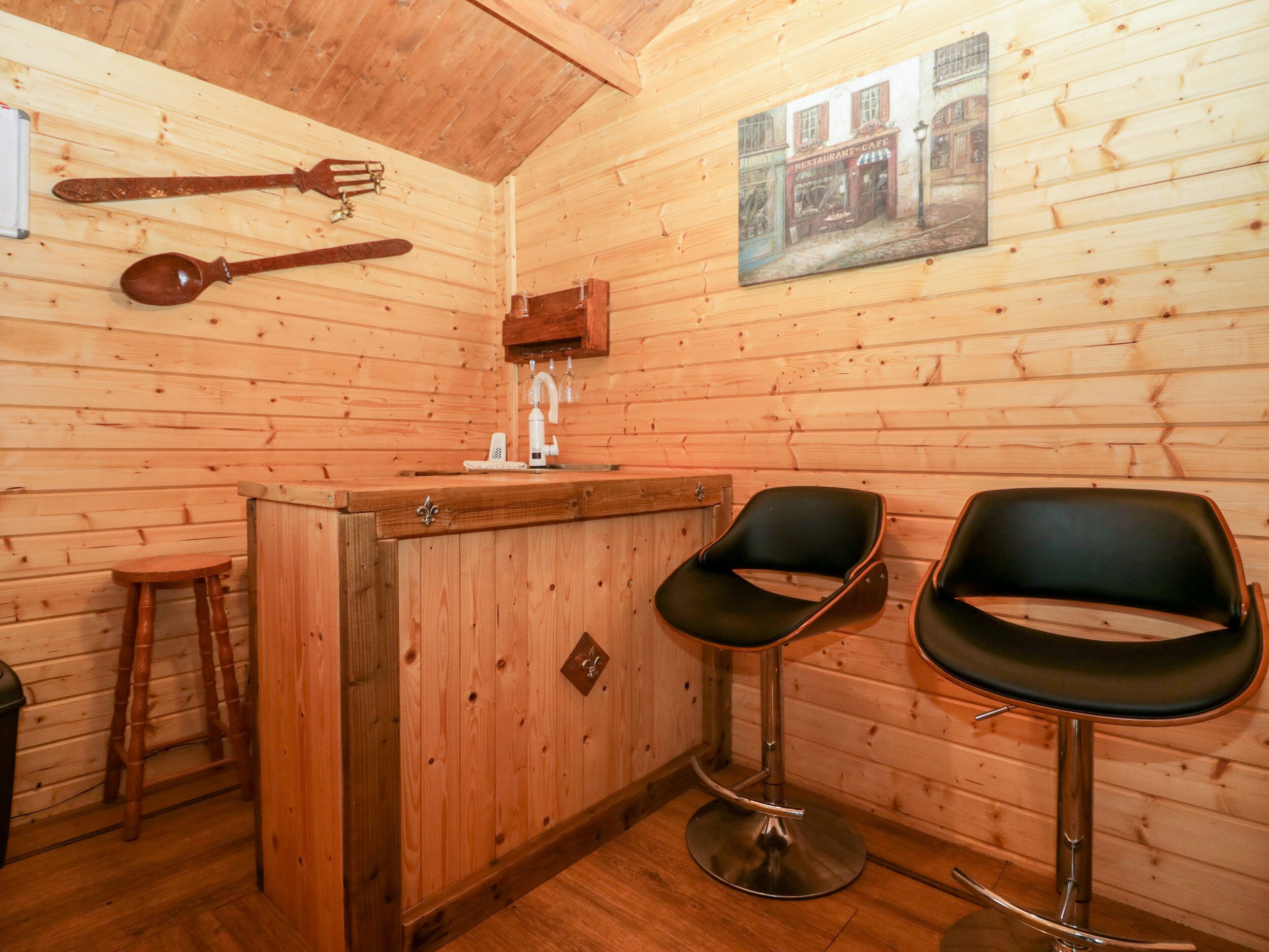 Een houten interieur met een bar, twee zwarte barkrukken, een houten kruk en een houten wand met een afbeelding.