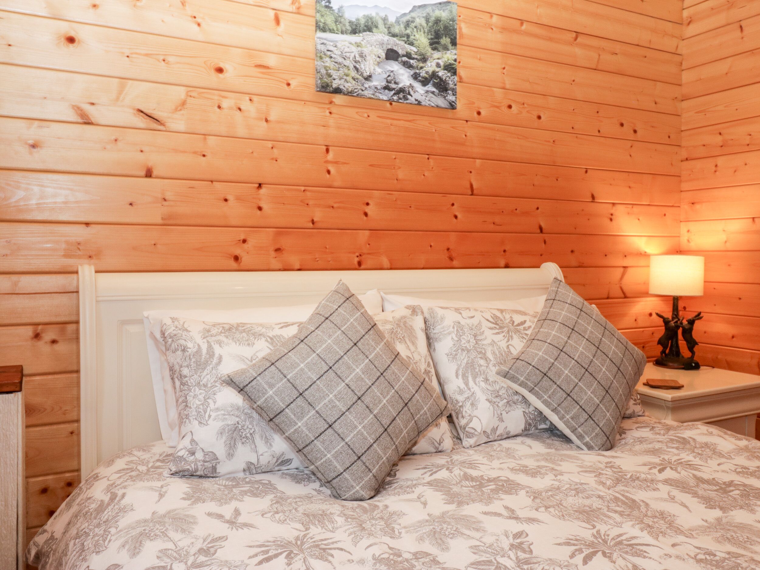 Una habitación de cabaña de madera con una cama, dos almohadas, una mesita de noche con una lámpara y un cuadro en la pared.