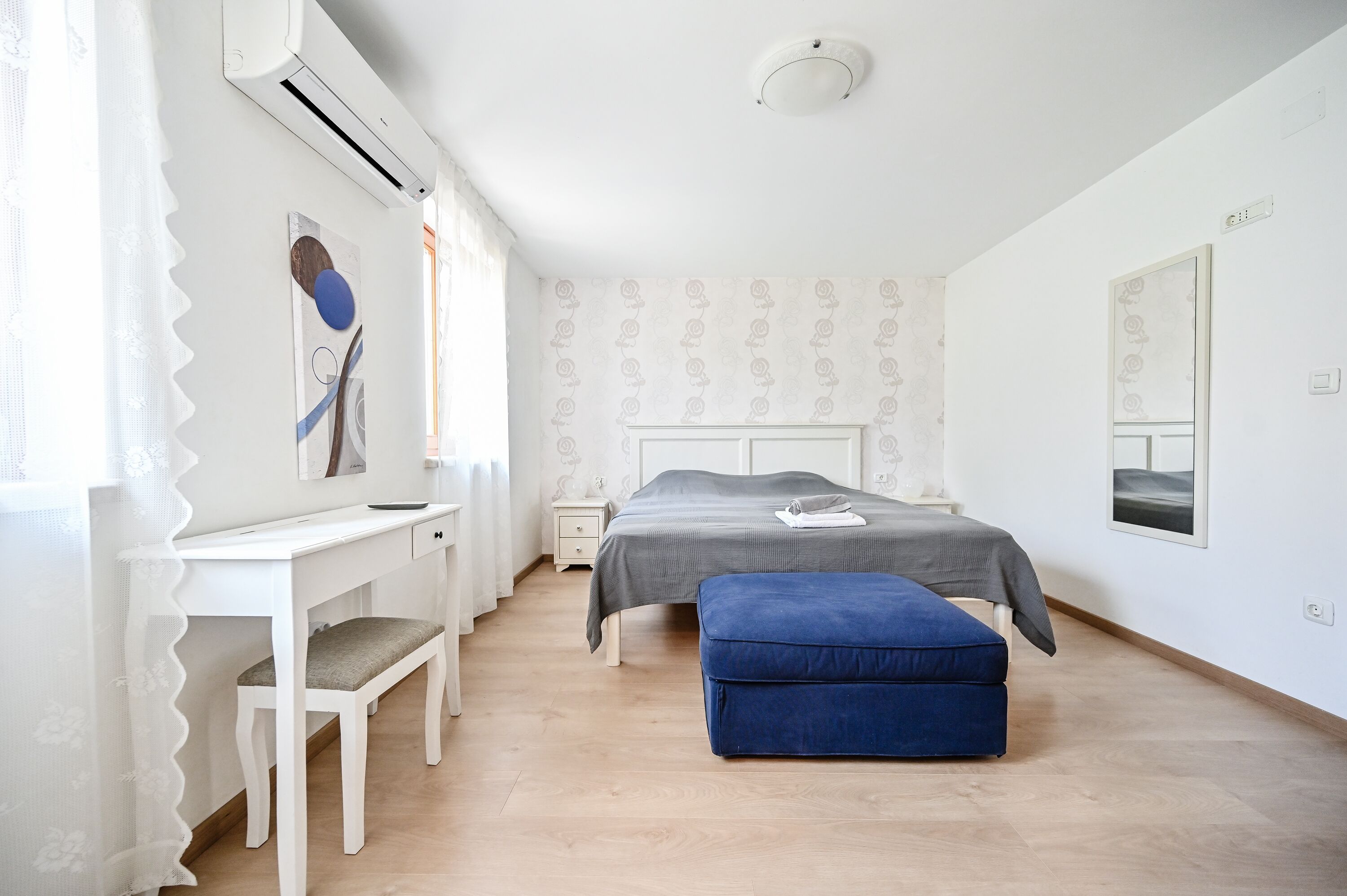 A bedroom with a bed, a desk, a chair, a bedside table, and a window with curtains.
