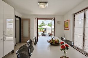 A dining area with a long table, chairs, a fruit bowl, and a vase with flowers.