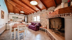 A rustic kitchen with a stone wall, wooden ceiling beams, a fireplace, and a dining area with a table and chairs.